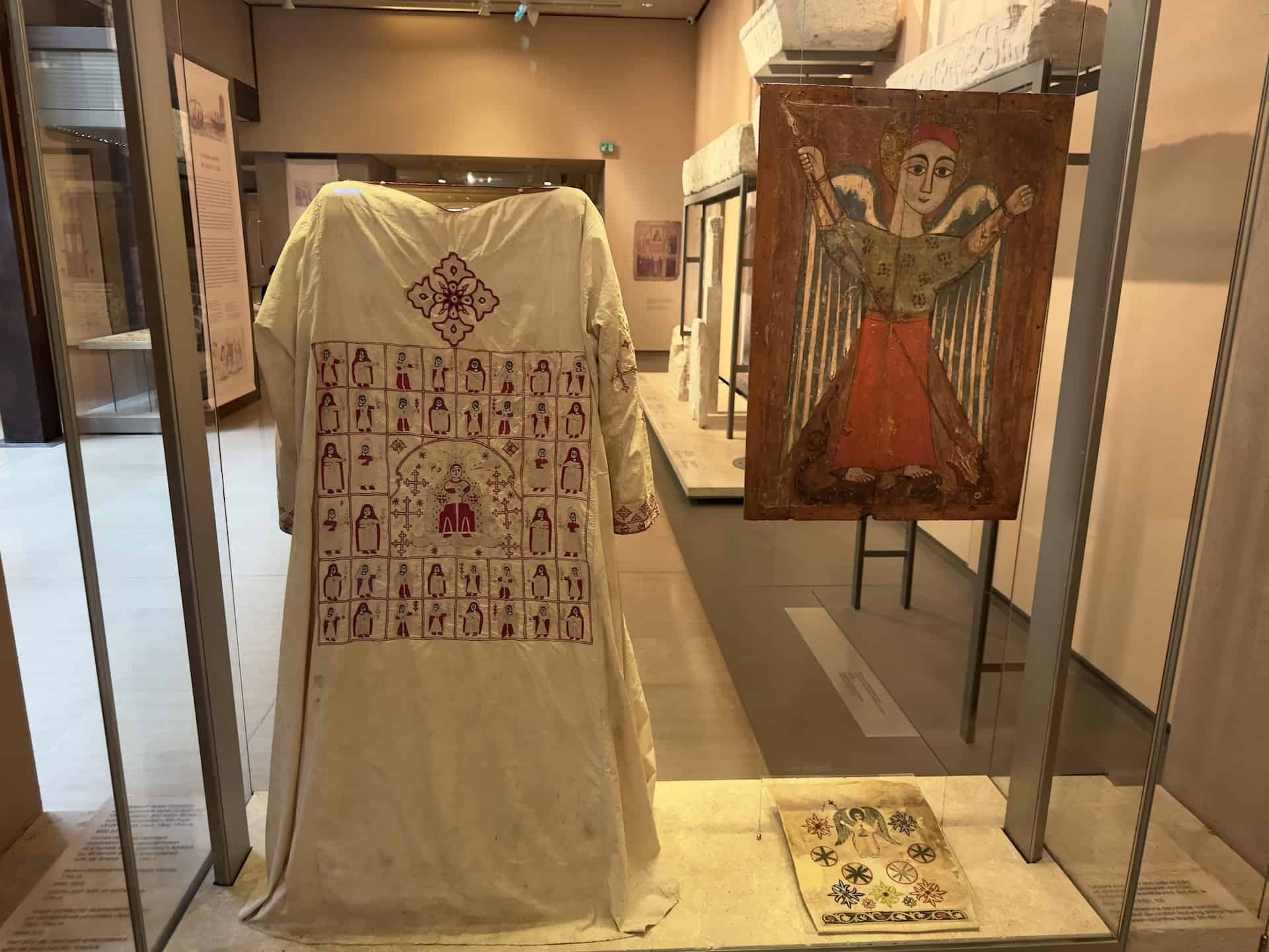 Cotton liturgical vestment (sticharion) with representation of a female saint (Damiana?) flanked by 40 female saints, embroidered with silk thread, 18th-19th century (left); Coptic icon with an archangel, 17th century (top right); part of a silk vestment embroidered with silk and metallic threads, 15th-19th century (bottom right) at the Byzantine Museum in Athens, Greece